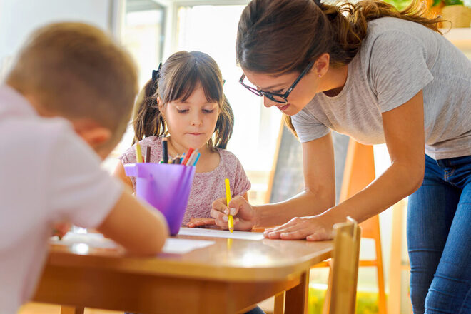 Kinder zeichnen an einem Tisch, eine Erzieherin unterstützt sie dabei