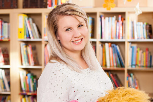 Eine blonde Frau mit weißem Tshirt steht vor einem Bücherregal mit Kinderbüchern. Sie hält eine Stoffpuppe in der Hand.
