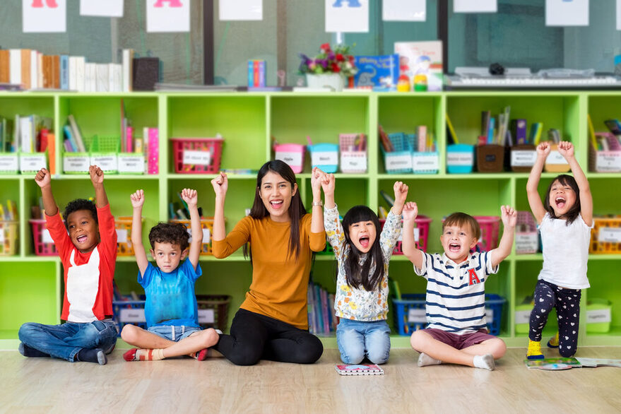 Eine junge asiatische Frau und fünf Kinder knien auf dem Boden und heben ihre Arme freudig in die Luft