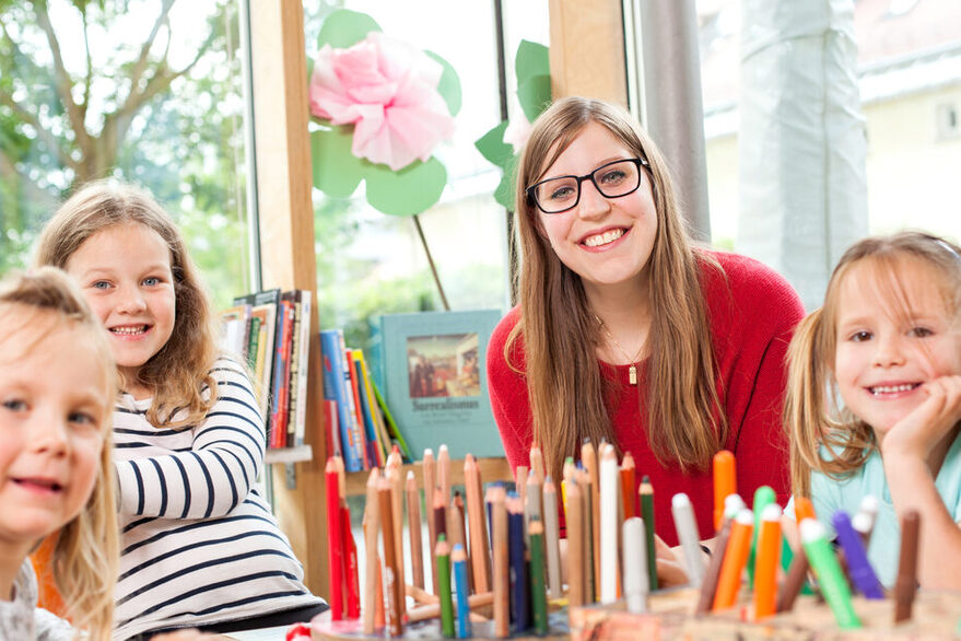 Erzieherin sitzt mit Kindern an einem Tisch voller Buntstifte