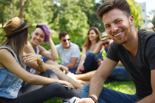 Eine Gruppe junger Leute sitzt auf einer Wiese, der junge Mann im Vordergrund lächelt in die Kamera
