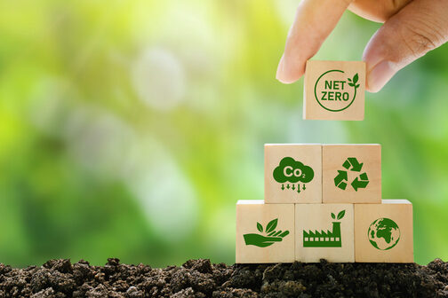Eine Hand legt einen kleinen Holzwürfel mit der Aufschrift "Net Zero" auf eine Pyramide aus weiteren Würfeln, auf denen Piktogramme zum Thema Nachhaltigkeit abgebildet sind.